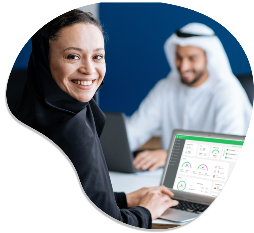 Woman in hijab smiling while working on a laptop, with a man in a thobe in the background